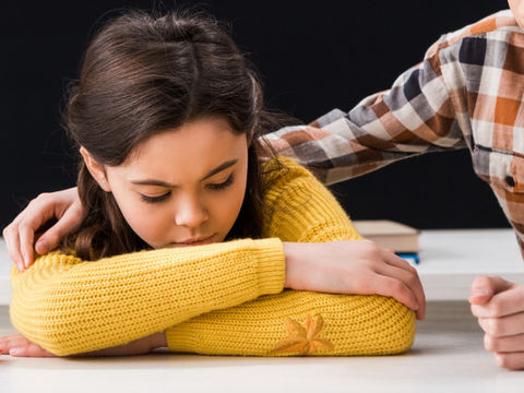 A girl with her head down on a desk, with an arm around her shoulder
