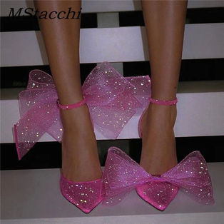 Sparkly pink high heels with large bows on a striped black and white background. Text reads "MStacchi" in the top left corner.