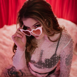 Woman in sheer top poses with pink heart-shaped sunglasses on, set against red curtains and a fluffy surface, creating a playful mood.