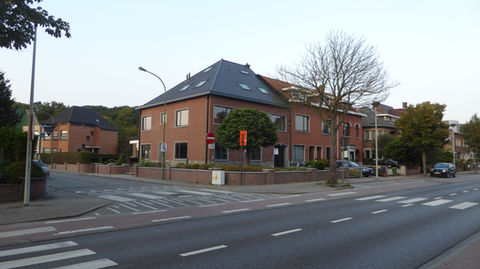 Hoekgebouw met roodbruine bakstenen gevel aan een brede straat, omringd door bomen en woonhuizen.