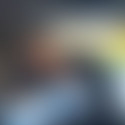 Smiling woman in a black jacket looks at a baby in a car seat inside a vehicle. Sunlit trees are visible outside the window.