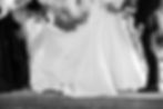 Bride in white gown holding bouquet, groom in suit beside her. Maid of honor adjusts train. Black and white photo, outdoor setting.