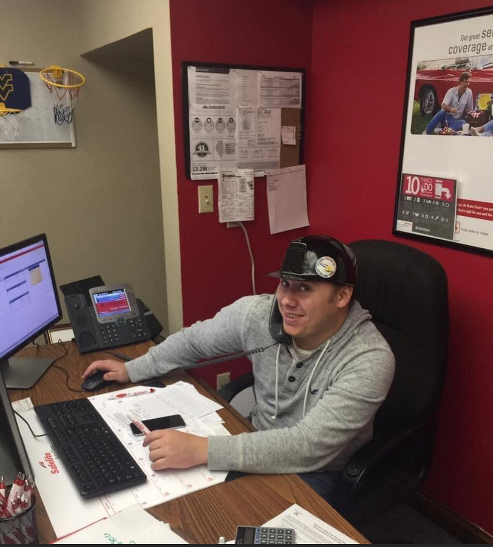 Eric Dotson multitasking at their office desk, wearing a hard hat while on the phone, surrounded by paperwork and a computer, with a playful basketball hoop on the wall for lighthearted moments.