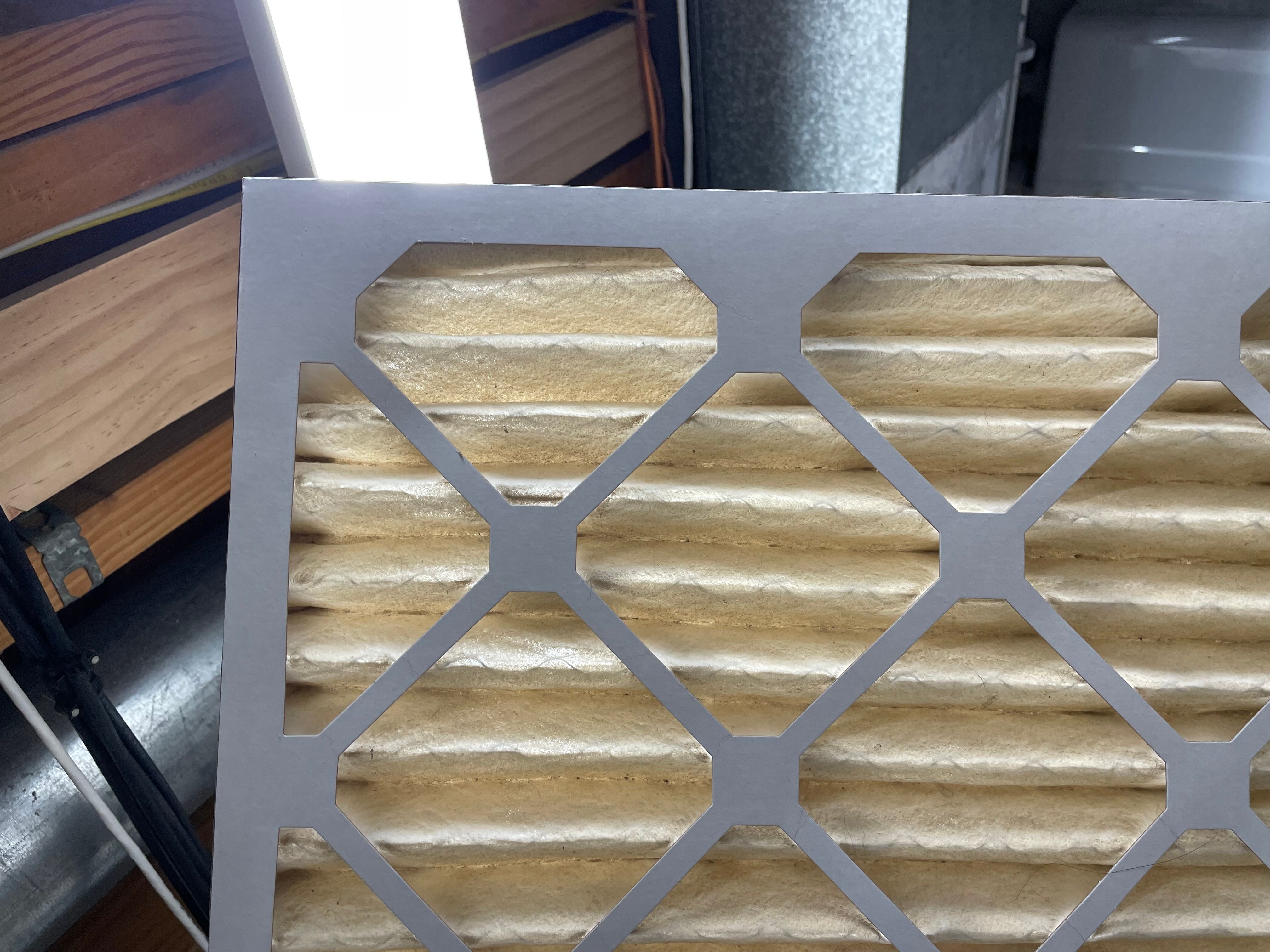 A used HVAC air filter is being held up in front of a long fluorescent light. The filter is noticeably dirty and very little light can be seen through it. 