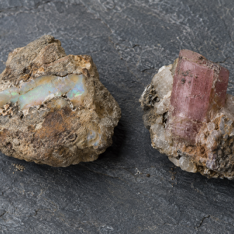 Two rough mineral rocks on dark slate: one with iridescent opal-like vein, the other with a pink tourmaline.