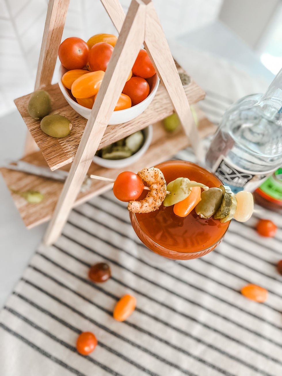 A Bloody Mary martini placed on a striped cloth with a vegetable and shrimp skewer placed across the top of the glass as a garnish. Behind the glass is a wooden 3 tiered tray with bowls of tomatoes, pickles and olives placed on it.