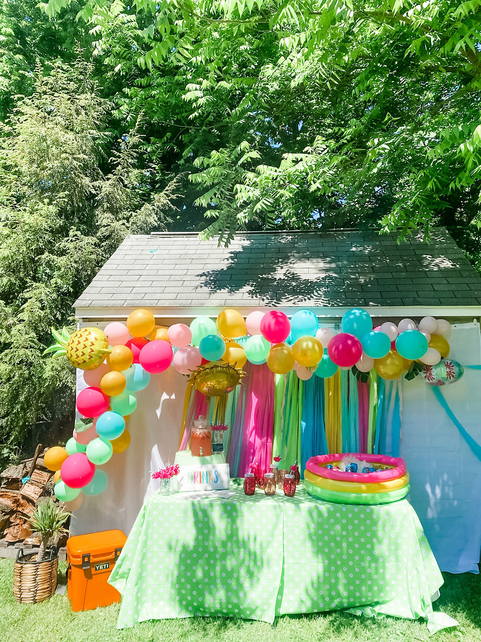 A drink station with a lime green polka dot cover, a colorful blow up pool used as a cooler for drinks, Hippie Juice in a canister and pink flamingo cups. A streamer backdrop was used and a bright balloon arch with Bright pink, light pink, tiffany blue, yellow and green.
