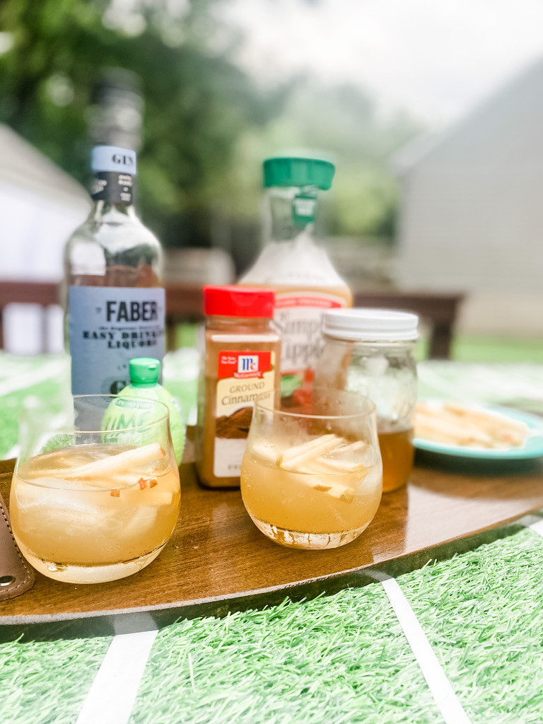 Two fluted rocks glasses filled with an Apple and Gin cocktail with the ingredients behind the cocktails. Faber gin, ground cinnamon, apple cider, honey simple syrup and apple straws all placed on a wooden cutting board outside.