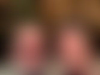 Two adults smiling at the camera in front of a decorated Christmas tree and warm string lights. One wears glasses and a gray patterned sweater with a beard, the other has a styled quiff, mustache, and a patterned sweater; the cozy background features gold ornaments and soft, glowing lights.