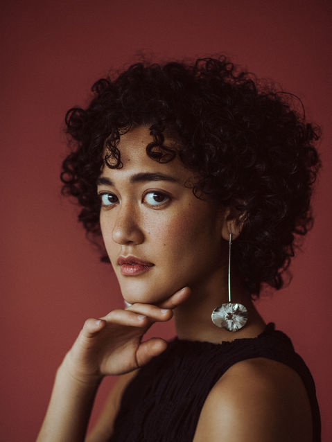 Model with short curly hair, wearing long silver earrings on a red backdrop.