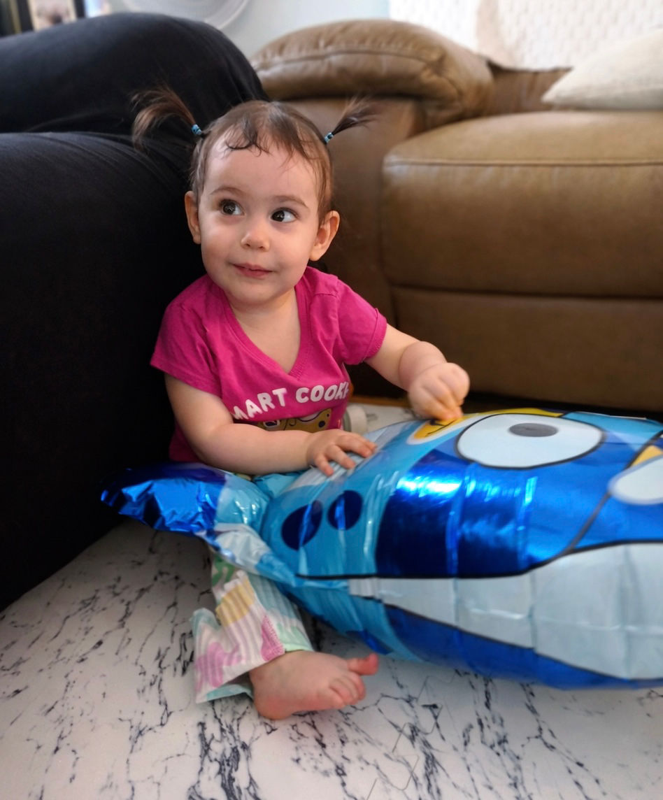 Ellie sits on the floor holding a shiny blue balloon, looking off to the side with a soft, curious expression.