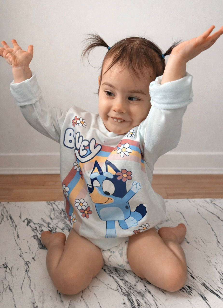 Ellie sits on the floor with her hair in two little pigtails, smiling with both hands raised in the air, wearing a Bluey shirt.