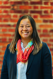 A smiling Southeast asian woman, looking into the camera with a blurred brick background. Long black, brown, and blonde highlighted hair, wearing a navy blazer, white shirt, and pink, green, blue scarf.
