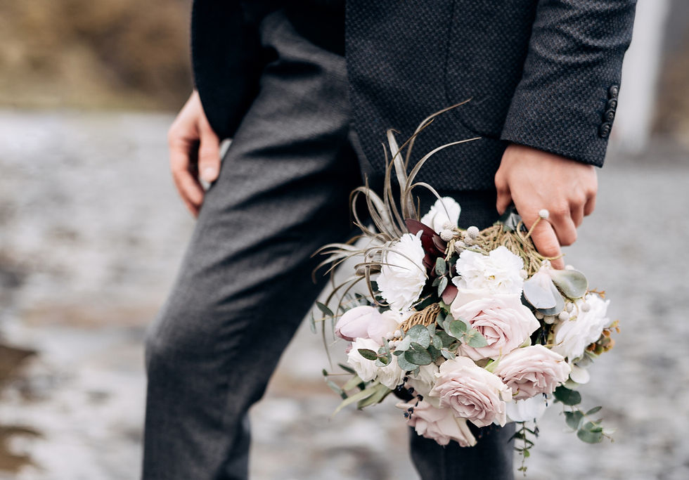 The lower torso and legs of a man in a suit holding flowers with them facing the ground