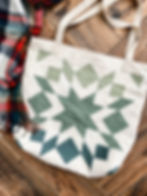 Cream tote bag with a star pattern in shades of green laying on a wood floor with a plaid scarf.
