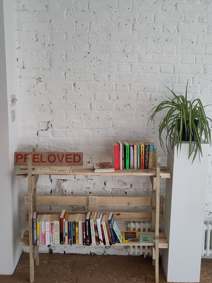 Bookshelf with books and a sign saying PRELOVED in red letters. A plant is on a white stand.