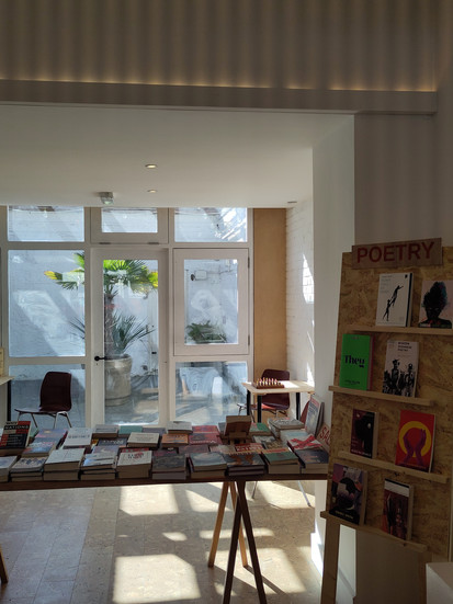Picture of large white room with big windows at the end letting in sunlight. The room is filled with bookshelves and a table filled with books. The mood is calm.