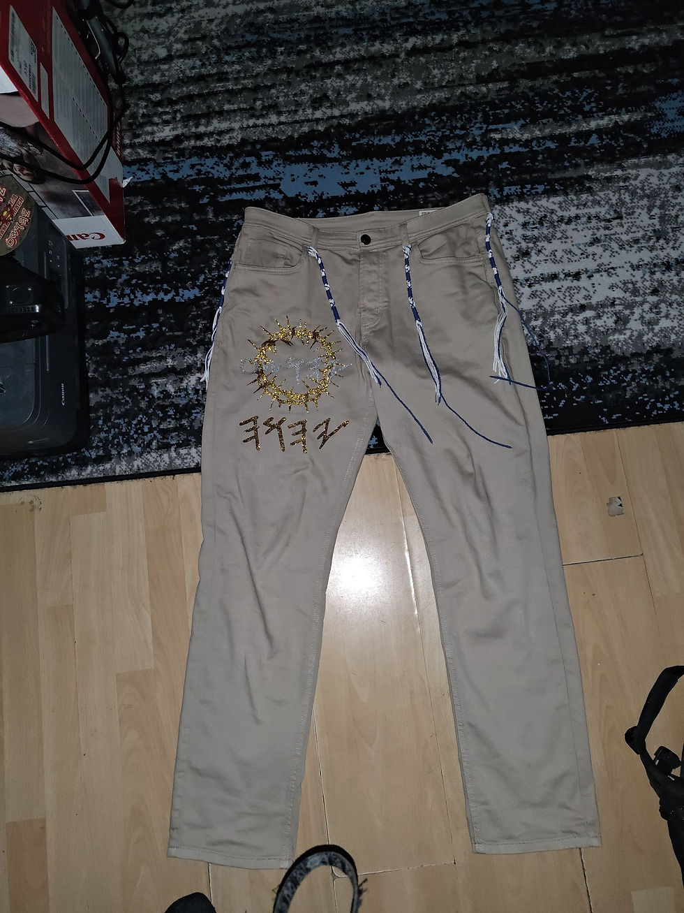 Thumbnail: Tan pants with art, text, and decorative strings on a wooden floor Yahusto.