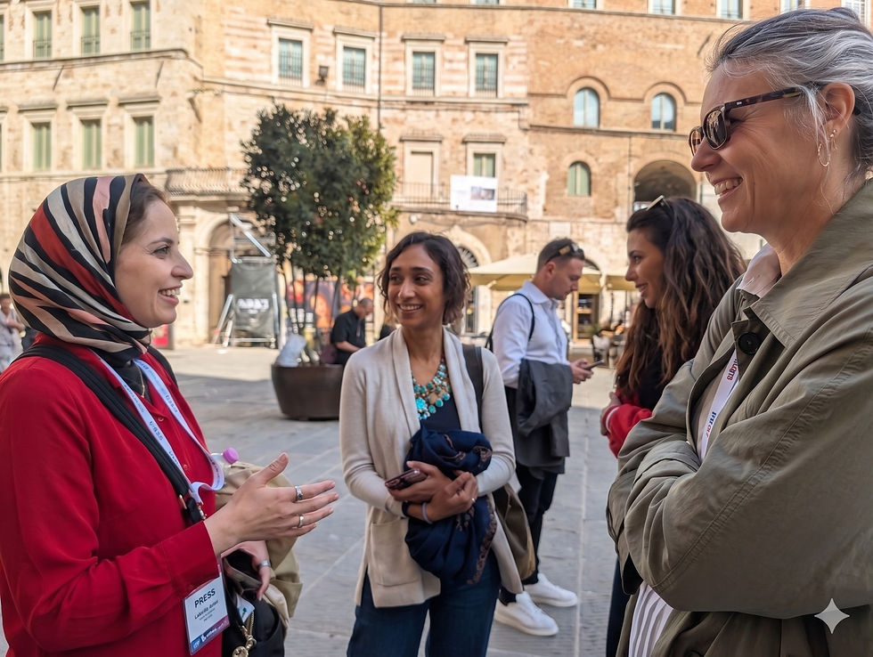 Converging in the Core: Journalists and documentarians Singeli Agnew (left) and Laila Al-Arian (center) are seen in deep conversation in the historic center of Perugia, Italy, during the 2026 International Journalism Festival. A prominent 'IJF Perugia 2026' festival banner is visible on the ancient stone facade, and Agnew wears her official festival credential, linking the professional community to the unique Italian hill town setting. Photo by Gretel Ehrlich.