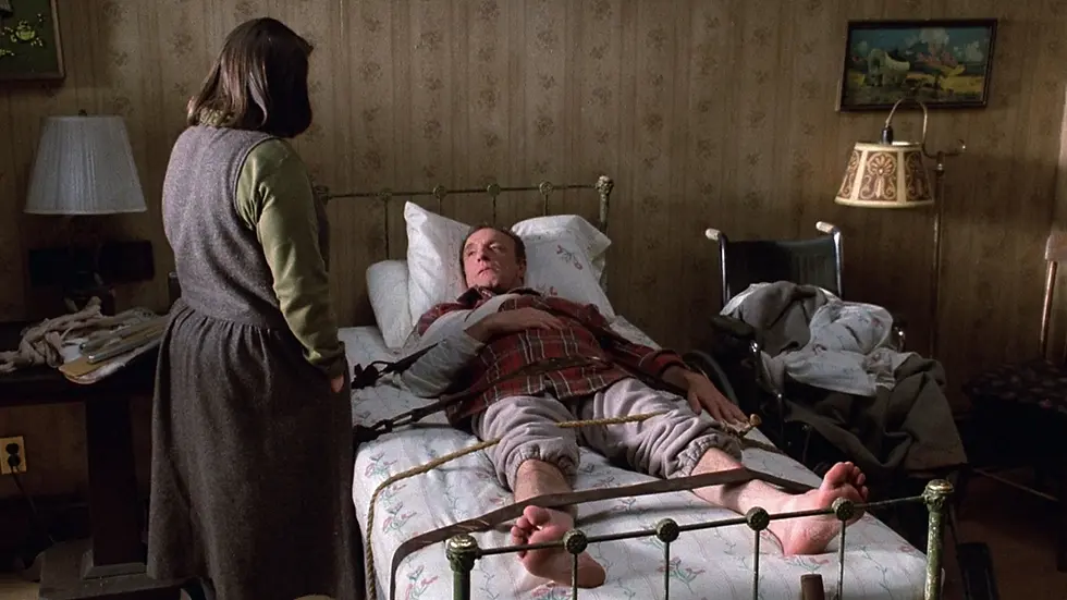 A person in plaid shirt is tied to a bed, watched by someone standing. The setting is a dimly lit room with vintage decor. Tense mood.