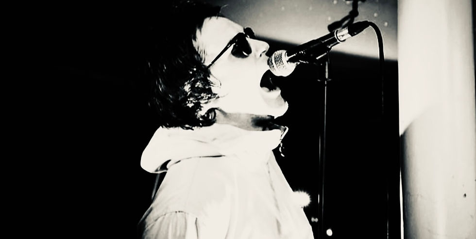 Singer in sunglasses passionately performs into a microphone in a dimly lit setting. Black and white tones enhance the dramatic mood.