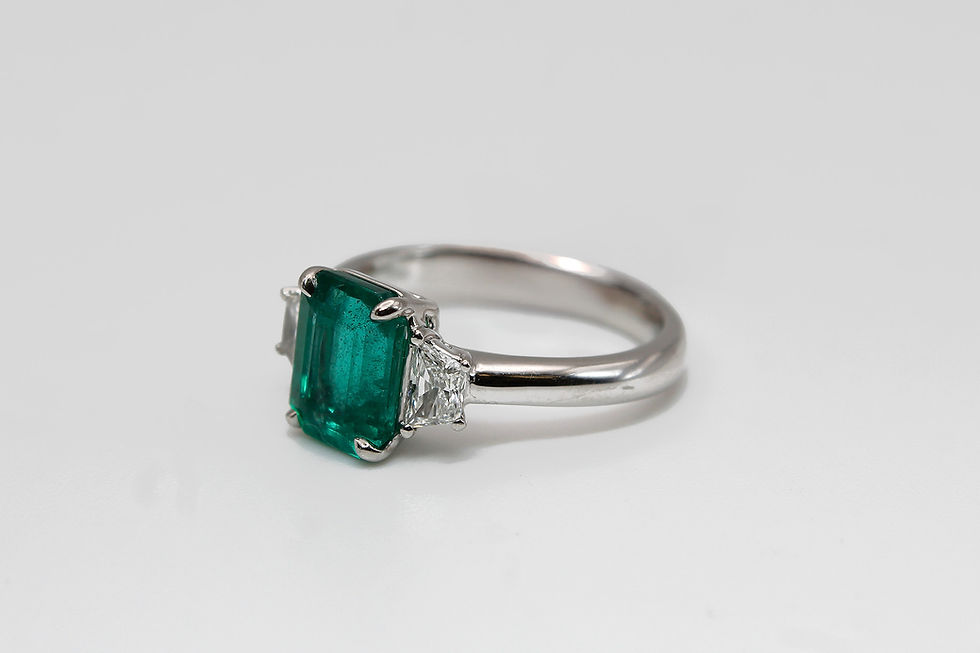 platinum ring with a large emerald center stone flanked by two triangular diamonds, set against a plain white background. Elegant and luxurious.