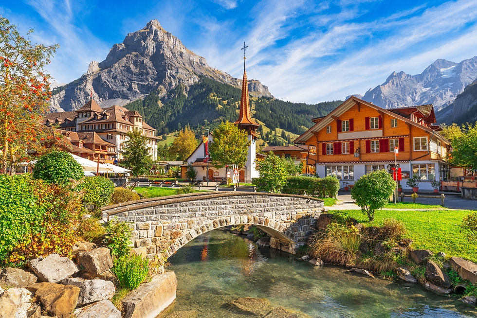 Charming village nestled in the Swiss Alps, showcasing picturesque wooden chalets, a quaint church with a tall steeple, and a serene stone bridge over a clear blue stream, all set against a backdrop of majestic mountains under a bright blue sky.