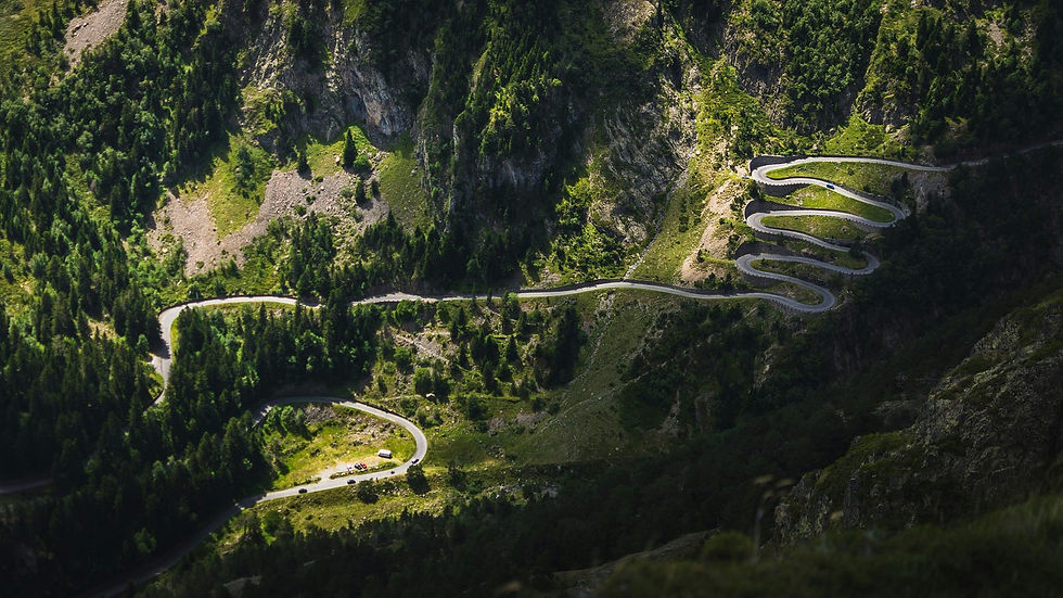 Aerial view of a winding mountain road snaking through lush, green forests and rugged terrain, showcasing a series of sharp hairpin turns.