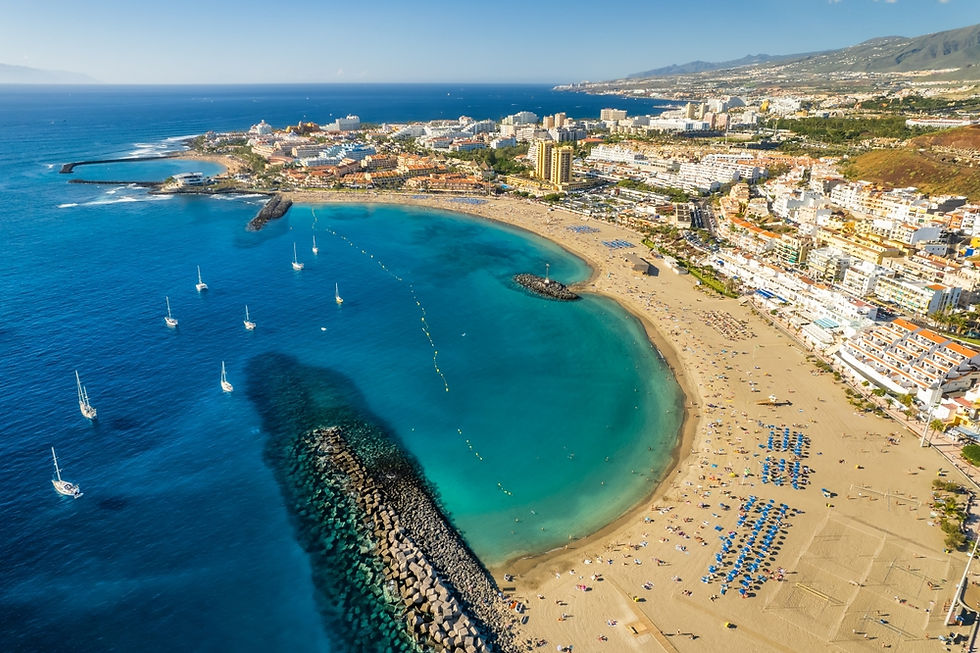 A stunning aerial view of a vibrant beach in Tenerife, where turquoise waters meet golden sands, dotted with sunbathers and surrounded by a lively coastal town. Sailboats gracefully float nearby, adding to the serene and picturesque setting.