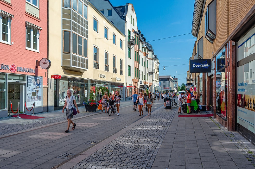 Shoppers stroll down a vibrant European street lined with colorful buildings, enjoying the sunny day and window shopping at popular stores.
