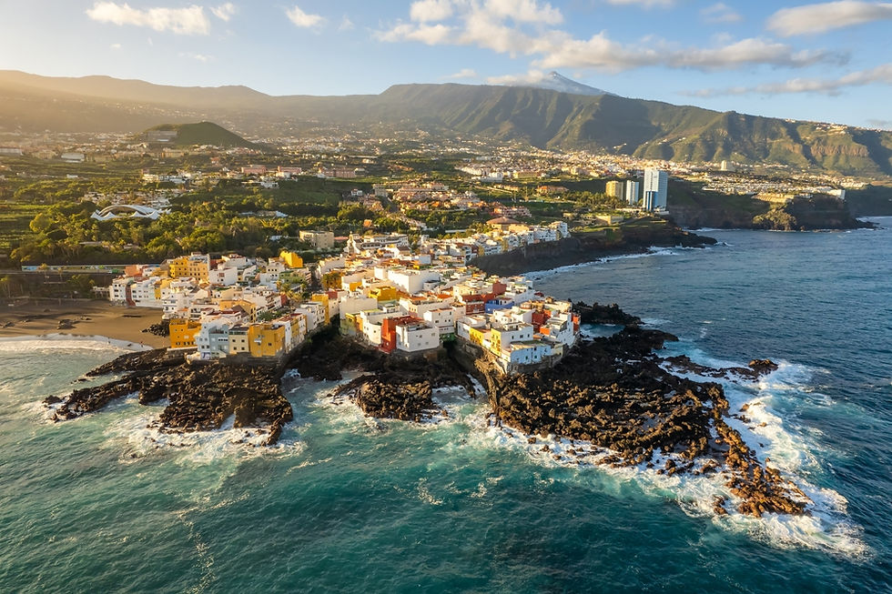 A breathtaking aerial view of a coastal town on Tenerife, Canary Islands, with vibrant houses perched on rocky cliffs, lush green landscapes, and the majestic Mount Teide in the background under a bright, clear sky.