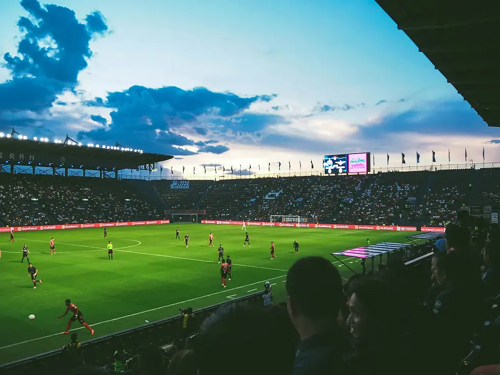 A lively evening soccer match unfolds in a bustling stadium, with vibrant green turf contrasting against the dramatic sunset sky. Spectators are engrossed in the action as players chase the ball under shining stadium lights.