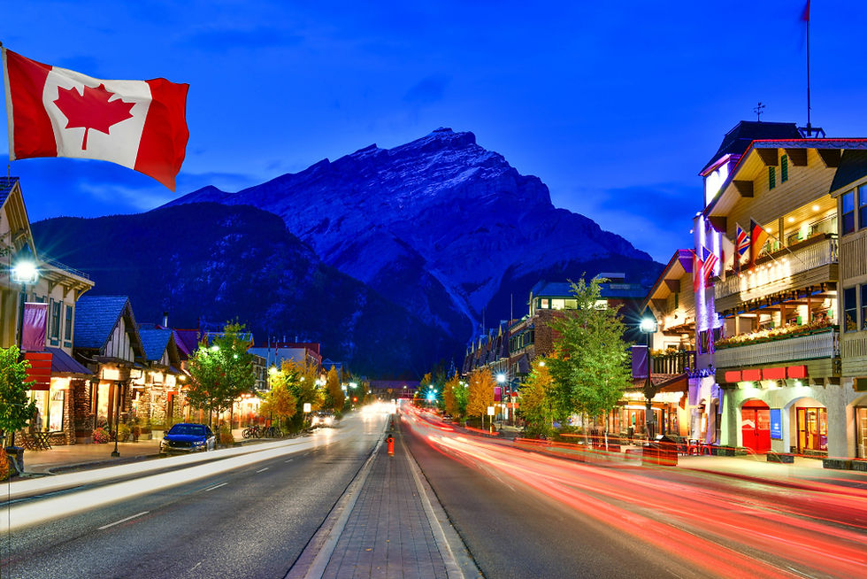 Vibrant evening lights illuminate the charming main street of Banff, Canada, as the Canadian flag flutters against a majestic mountainous backdrop.