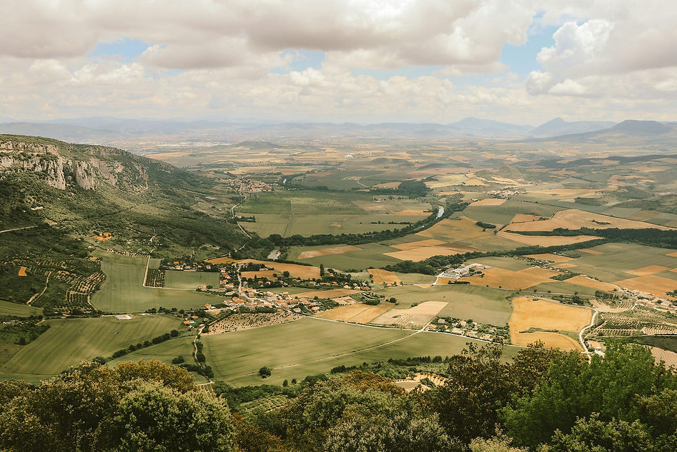 Expansive view of a rural landscape showcasing lush green fields, patchwork farmland, and a small village nestled at the foot of rolling hills under a partly cloudy sky.