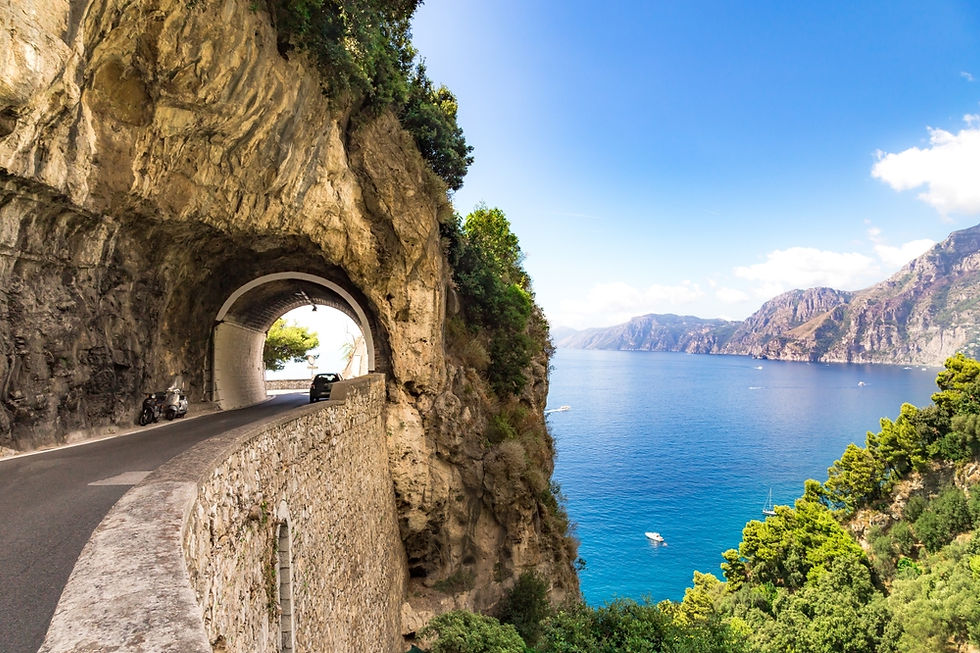 A scenic coastal road winds through a rocky tunnel along the stunning Amalfi Coast, offering dramatic views of the azure sea and rugged cliffs under a bright blue sky.