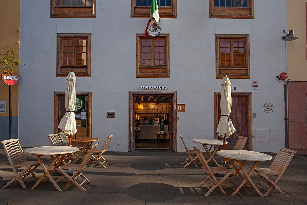 Charming façade of a Starbucks nestled in a historic building, featuring outdoor seating with umbrella-shaded tables, framed by rustic wooden windows and adorned with a national flag overhead.
