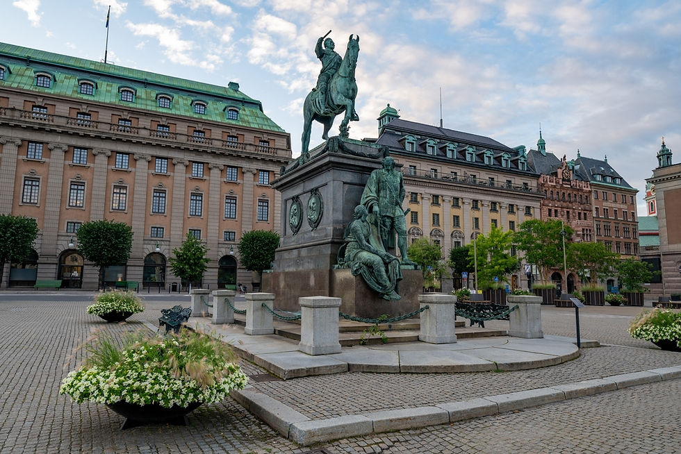 A majestic equestrian statue dominates the cobblestone square next to a historic building with a distinctive green roof, bordered by lush greenery and vibrant floral arrangements under a partly cloudy sky.