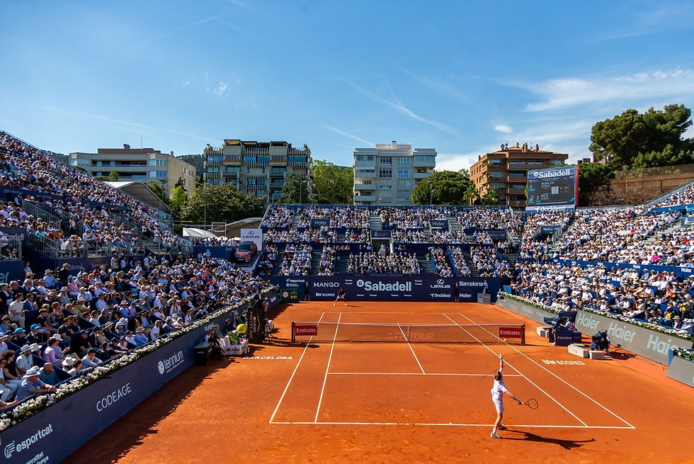 A packed crowd gathers under a clear blue sky at the Barcelona Open Banc Sabadell, as a player serves on the vibrant clay court surrounded by the stunning backdrop of the city.