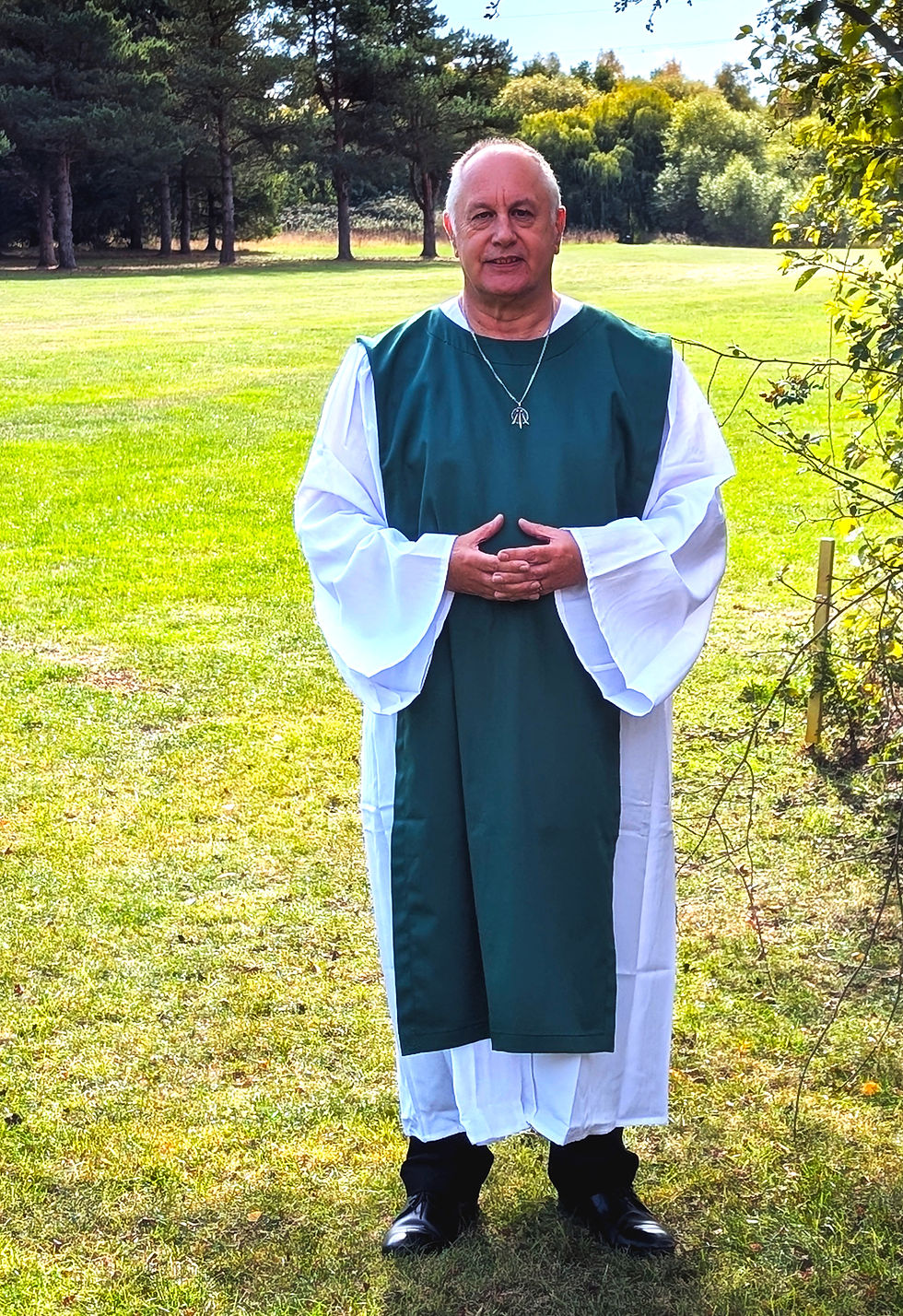A man dressed in celebrant robes posing outdoors on a sunny day with a rich green grass and trees around him