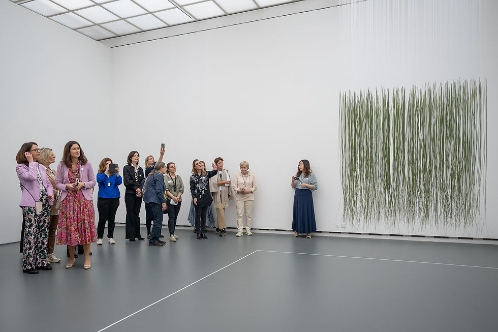 Un groupe d'environ 15 personnes, principalement des femmes, se tient dans un grand espace clair avec un mur blanc et un sol gris. Elles regardent une installation minimaliste sur le mur, composée de nombreux fils ou bâtons verts suspendus verticalement qui tombent du plafond. Plusieurs personnes dans le groupe tiennent des téléphones portables levés pour prendre des photos.