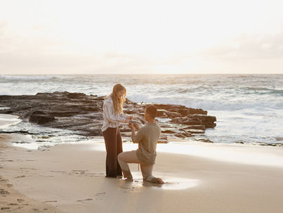 Man kneels proposing to a smiling woman on a sandy beach at sunset, waves crashing in the background. Romantic and serene setting.