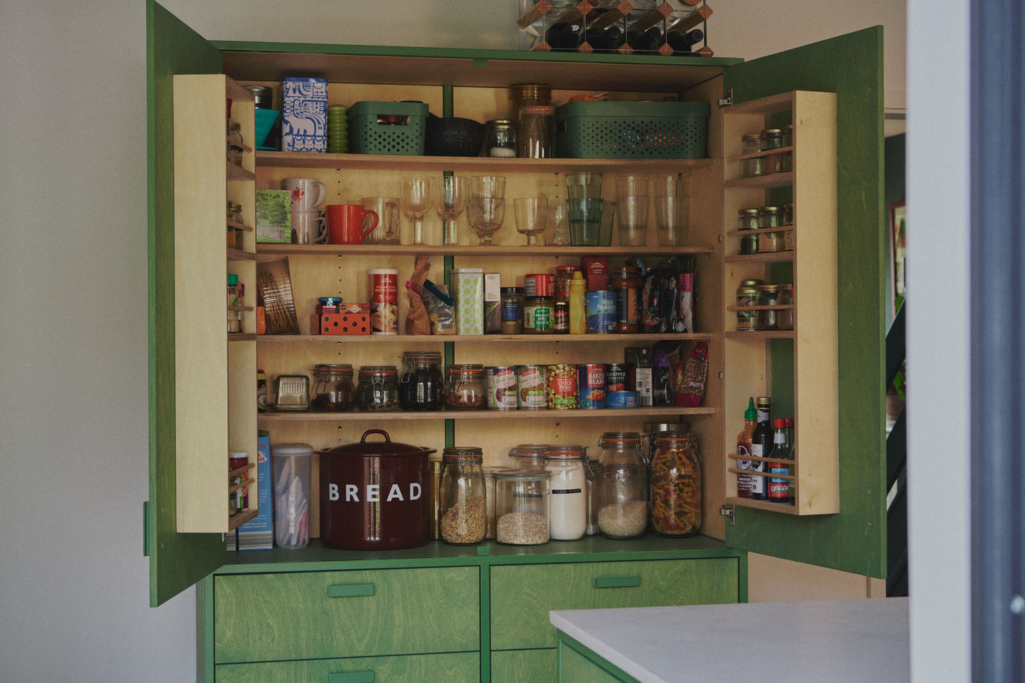 Sage green galley kitchen with RAL 6011 steel frames, stained birch plywood fronts, raw white quartz worktop, open shelving, and quarry tile floor.