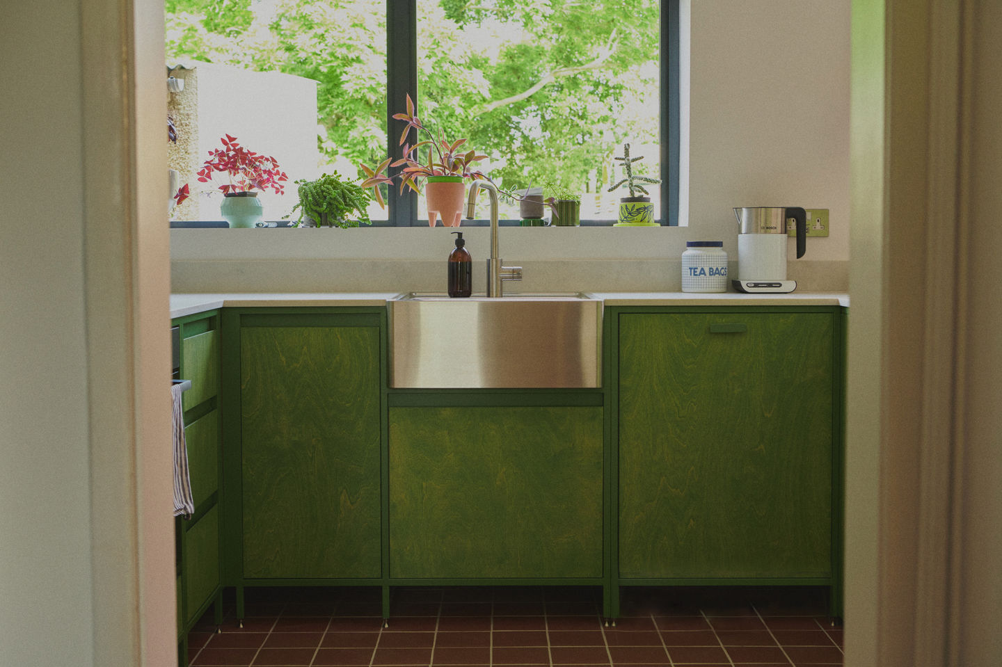 Sage green galley kitchen with RAL 6011 steel frames, stained birch plywood fronts, raw white quartz worktop, open shelving, and quarry tile floor.