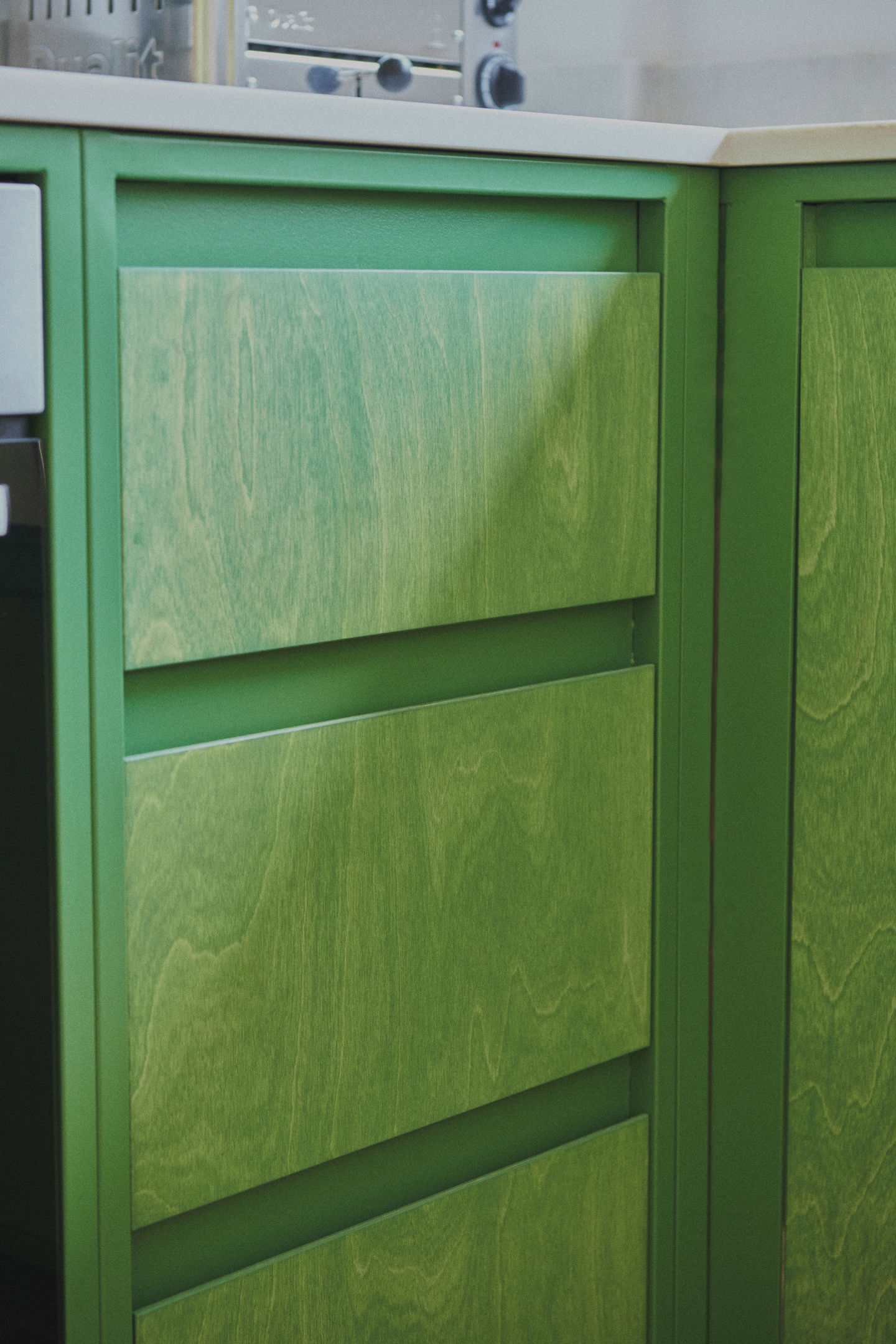 Sage green galley kitchen with RAL 6011 steel frames, stained birch plywood fronts, raw white quartz worktop, open shelving, and quarry tile floor.