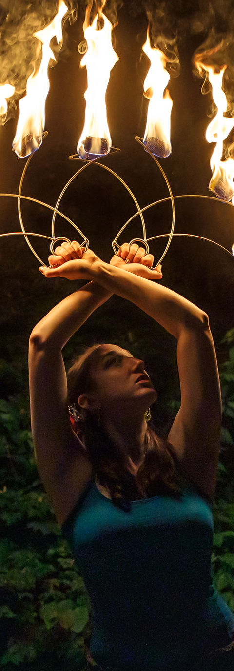 A fire performer holds flaming metal fans above her had at night, against a backdrop of trees.