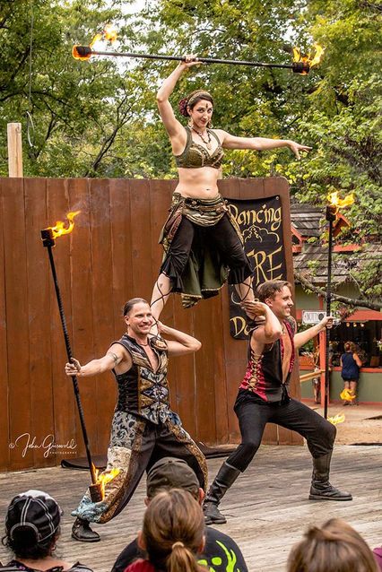 A female fire performer stands on the shoulders of two male fire performers, all holding fire staffs, on an outdoor stage