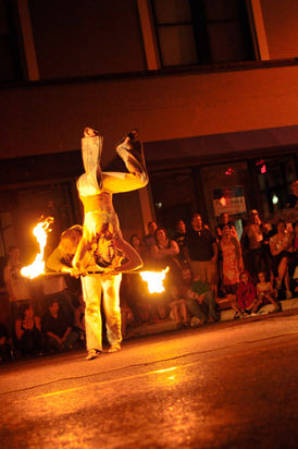 A female fire performer in a white costume flips over the back of a male fire performer holding a fire staff, in front of an audience