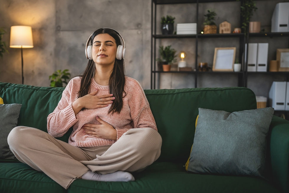Young woman sitting on a couch with headphones on, eyes closed, one hand on her chest and one on her belly, practising calming breath to soothe a panic attack.
