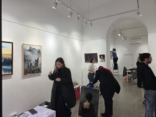 People in an art gallery hang paintings on white walls; one stands on a ladder. Art pieces depict landscapes and portraits. Calm atmosphere.