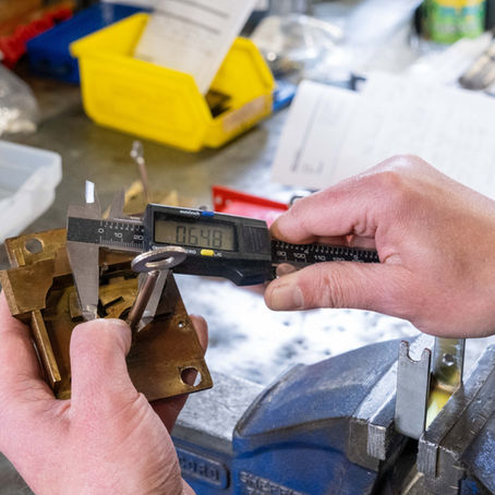 Hands use calipers to measure a metal lock part, reading 0.648 on a digital display. Workbench with tools and papers in background.